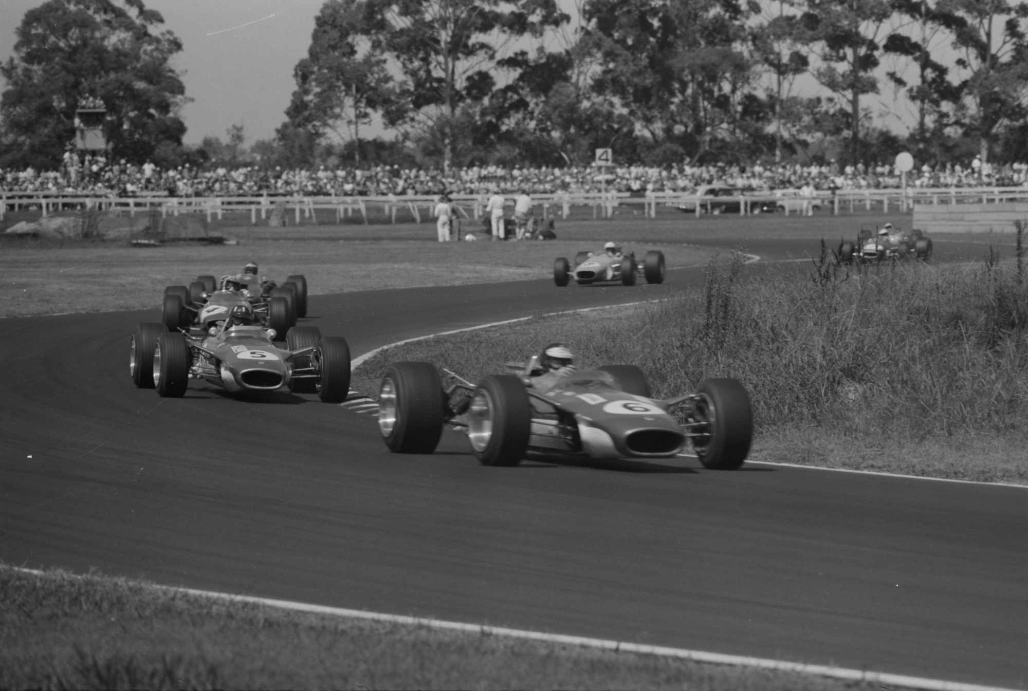 Revised alt text: Black-and-white photograph of Jim Clark, driving the number 6 Lotus, leading team-mate Graham Hill in the number 5 Lotus through a sweeping left-hand bend at Warwick Farm during the 1968 Tasman Series. Four other open-wheel racing cars follow, with a large crowd of spectators watching from behind wooden barriers and eucalyptus trees lining the circuit.